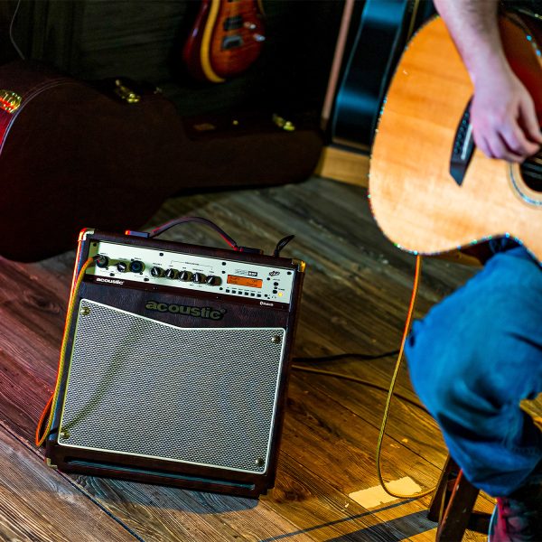 guitar player sitting in a stool performing with A40 40W acoustic amp, behind with guitars and guitar cases